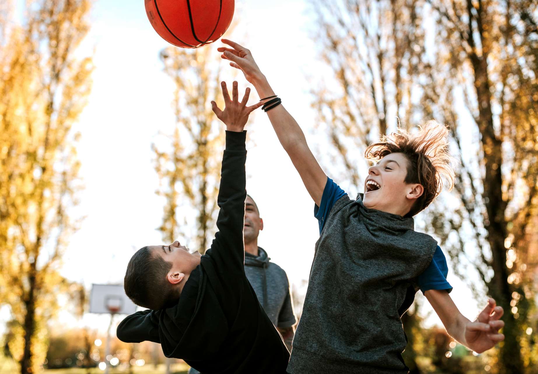 A candid photo of a father and his two sons play basketball at a local court in their neighbourhood. The two young boys are mid-jump trying to reach the ball.